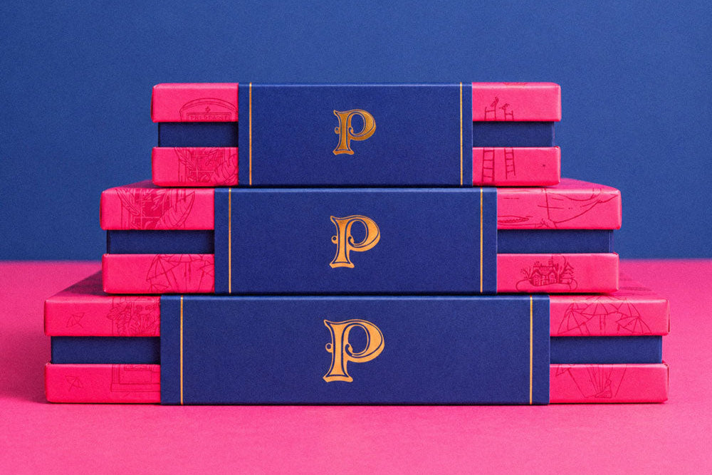 Three stacked rectangular prestat chocolate boxes or book-like packages on a pink surface against a blue background. The top and middle pieces are dark blue with a large gold letter P, and the side pieces are bright pink with a subtle map-like pattern.